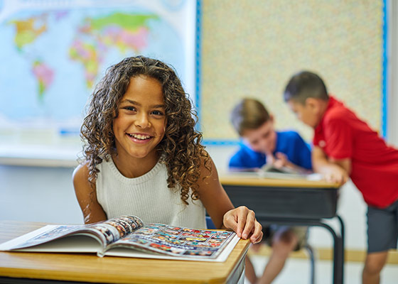 girl in the foreground with a yearbook looking at the camera and two boys in the background looking at a yearbook